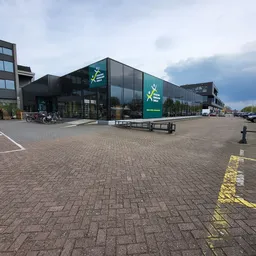 Voorgevel van kringloopwinkel met groene logo's en grote glazen pui aan een plein met parkeerplaatsen
