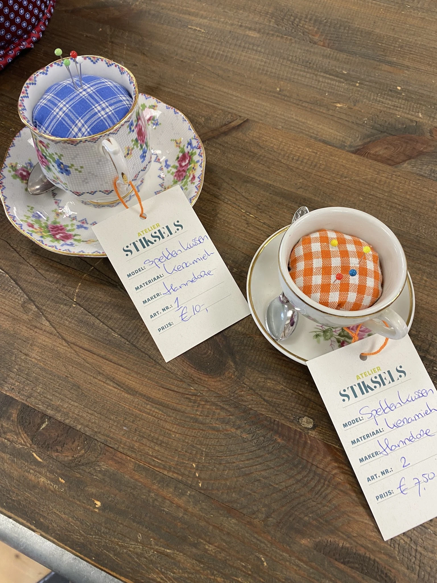 Twee porseleinen kopjes met schotels en STINSELS-labels, één met bloemenpatroon en één met oranje ruitpatroon.