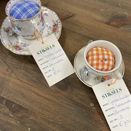 Twee porseleinen kopjes met schotels en STINSELS-labels, één met bloemenpatroon en één met oranje ruitpatroon.