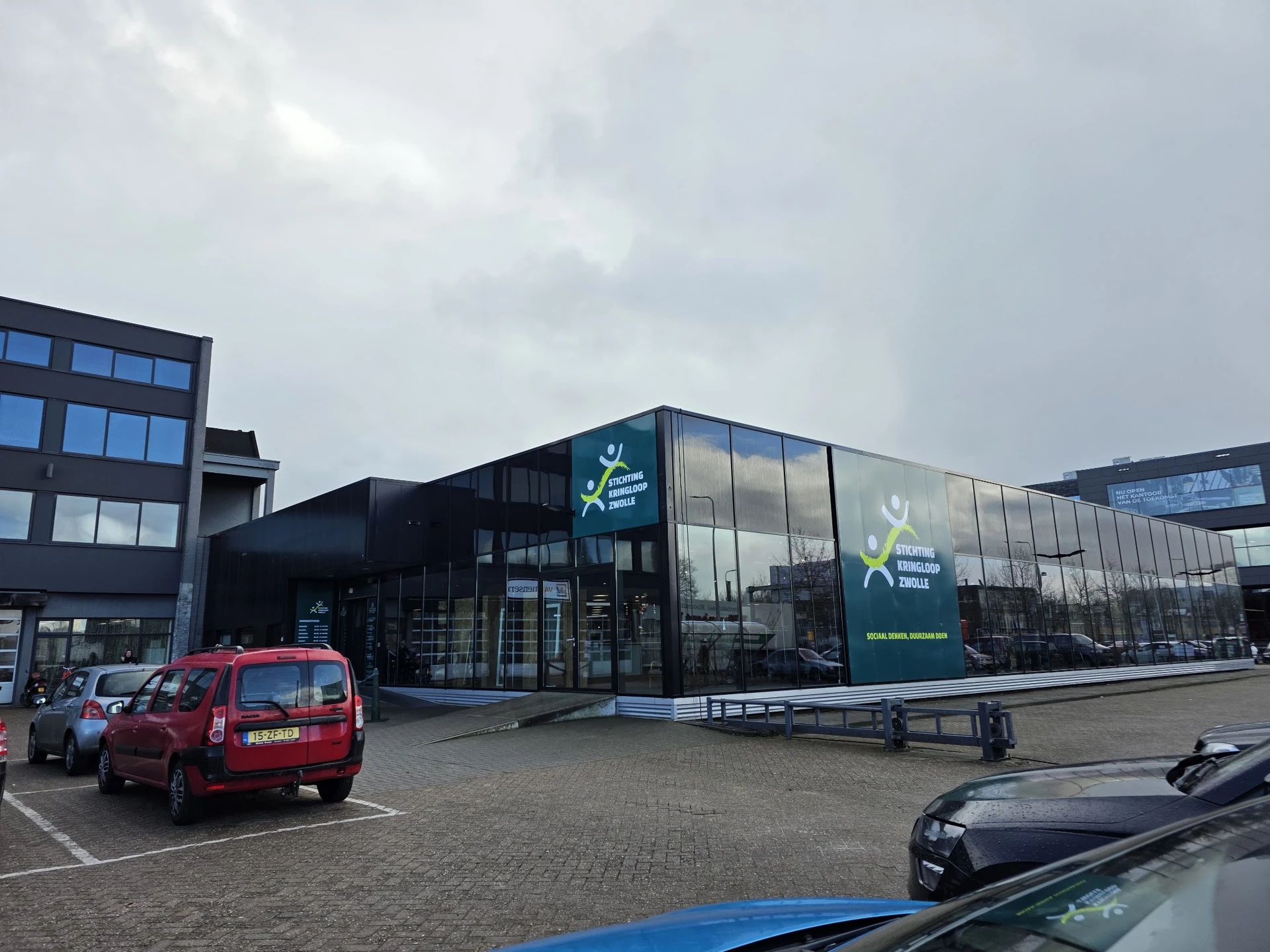 Moderne gevel van kringloopwinkel met grote ramen en groene branding, parkeerplaats met auto's ervoor