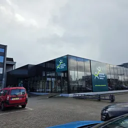 Moderne gevel van kringloopwinkel met grote ramen en groene branding, parkeerplaats met auto's ervoor