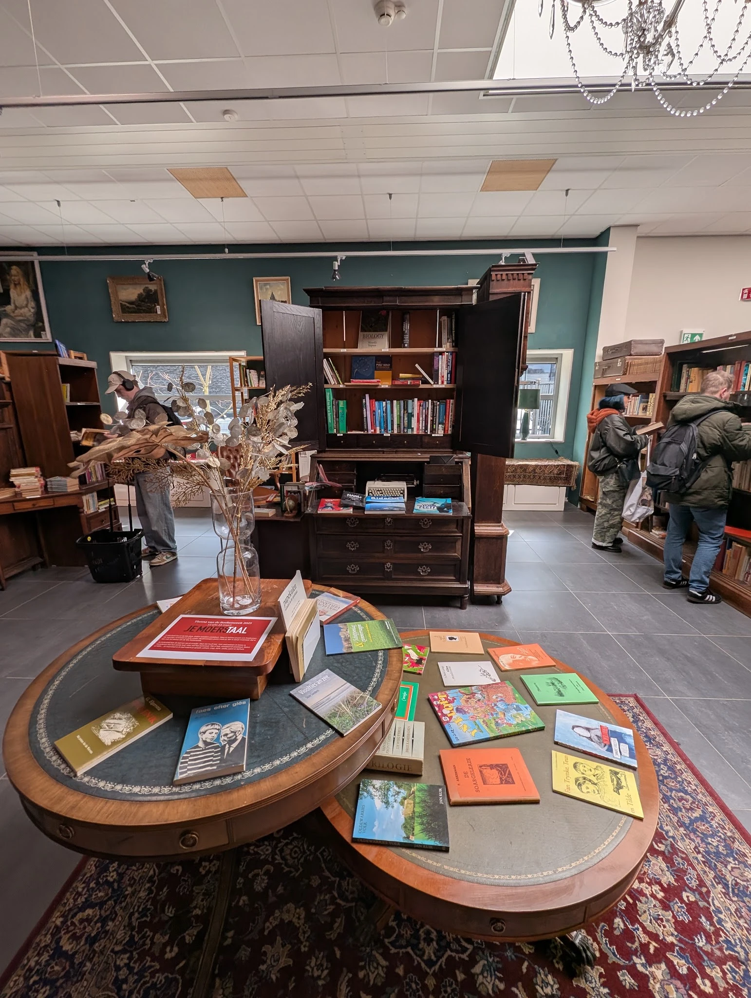 Boekenwinkelafdeling met houten boekenkasten, ronde tafel met boeken en gedroogde bloemen in een vaas, bezoekers in de winkel