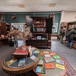 Boekenwinkelafdeling met houten boekenkasten, ronde tafel met boeken en gedroogde bloemen in een vaas, bezoekers in de winkel
