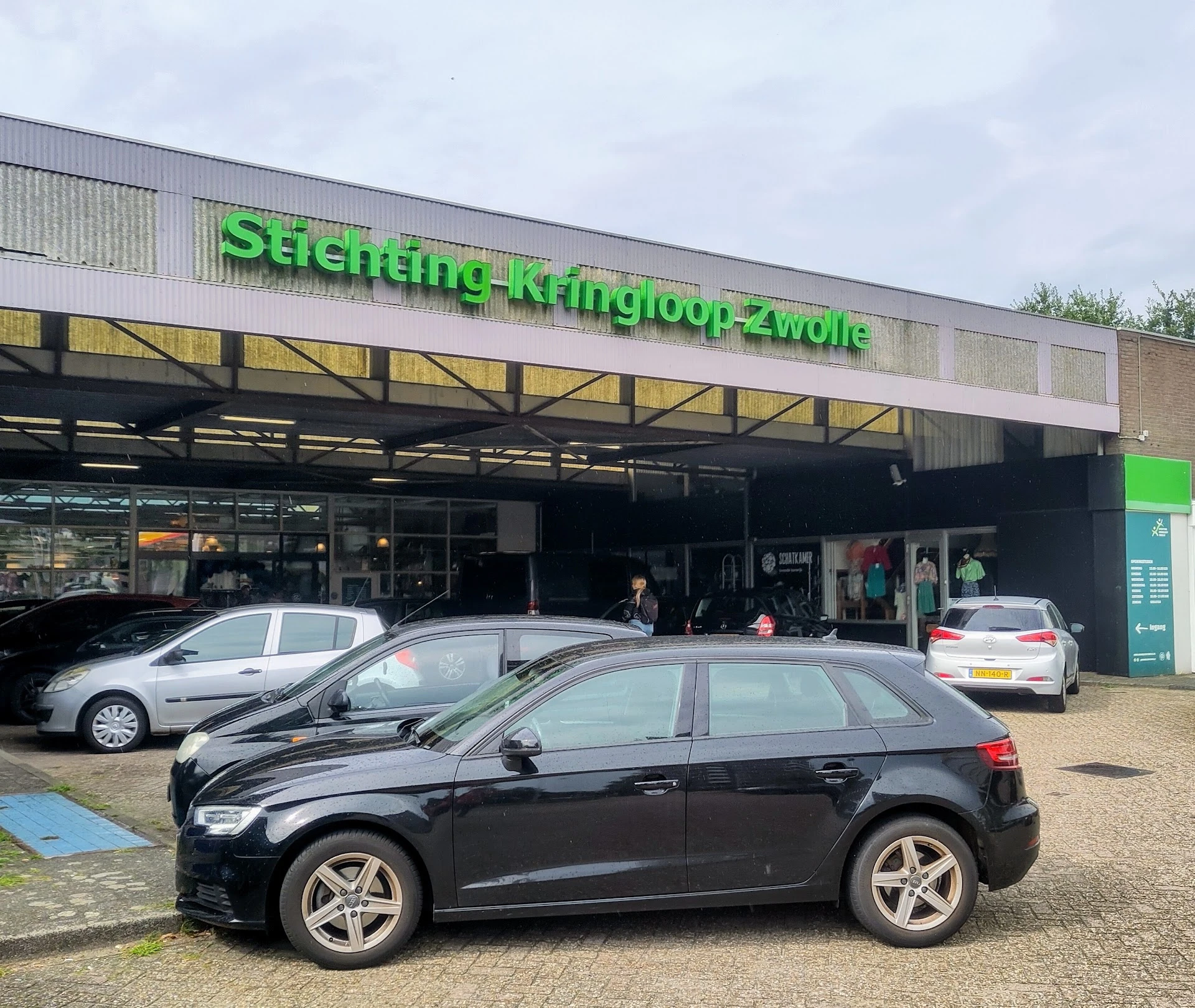 Voorgevel van kringloopwinkel met groene letters, parkeerplaats met auto's ervoor