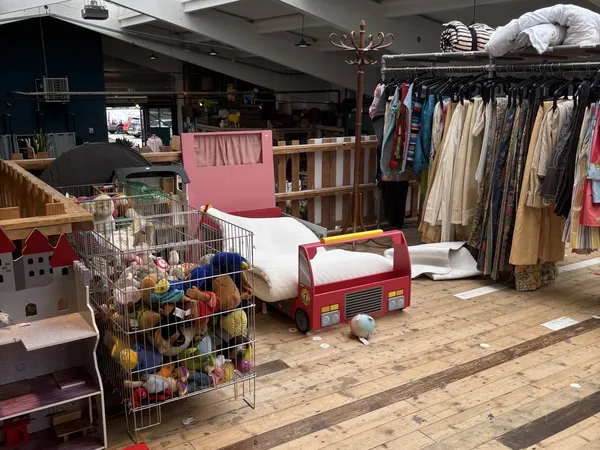 Speelgoedafdeling in kringloopwinkel met kinderbed, poppen, kleding op hangers en speelgoed in box