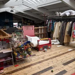 Speelgoedafdeling in kringloopwinkel met kinderbed, poppen, kleding op hangers en speelgoed in box