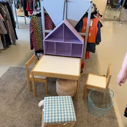 Kinderspeelgoed en meubels: houten tafel met stoelen, paarse speelhuisje, manden en gingham kussen in kringloopwinkel