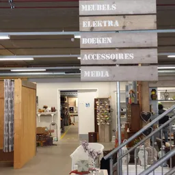 Winkelinterieur met houten hangborden met tekst 'ELEKTRA', 'BOEKEN', 'ACCESSOIRES', 'MEDIA', stellingen en diverse spullen zi