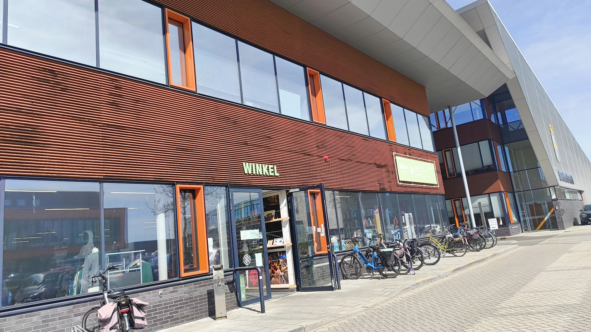 Gevel van kringloopwinkel met houten lambrisering, grote ramen en oranje details, met fietsen ervoor geparkeerd