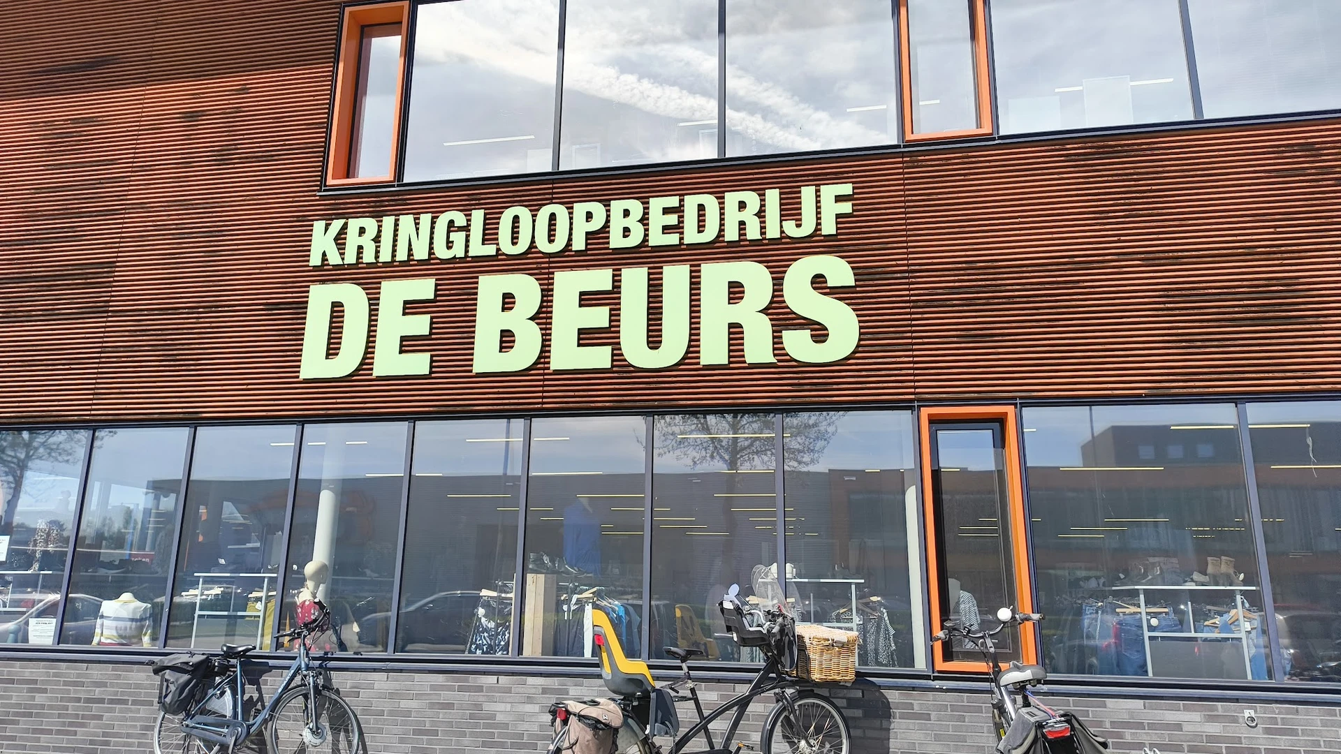 Gevel van kringloopwinkel De Beurs met grote gele letters op houten siding, grote ramen en fietsen geparkeerd ervoor