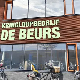 Gevel van kringloopwinkel De Beurs met grote gele letters op houten siding, grote ramen en fietsen geparkeerd ervoor