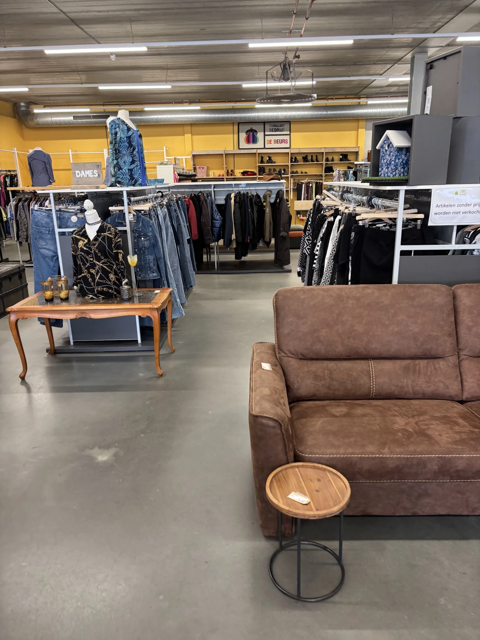 Winkelinterieur met bruine leren bank op voorgrond, kledingrekken met jassen en spullen op achtergrond, houten tafel met kled