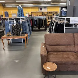 Winkelinterieur met bruine leren bank op voorgrond, kledingrekken met jassen en spullen op achtergrond, houten tafel met kled
