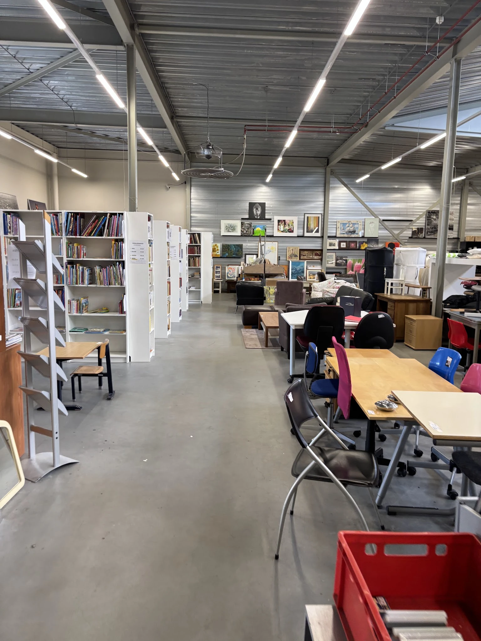 Winkelvloer met boekenkasten, tafels, stoelen en diverse tweedehands spullen in een ruim magazijnachtig interieur