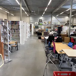 Winkelvloer met boekenkasten, tafels, stoelen en diverse tweedehands spullen in een ruim magazijnachtig interieur