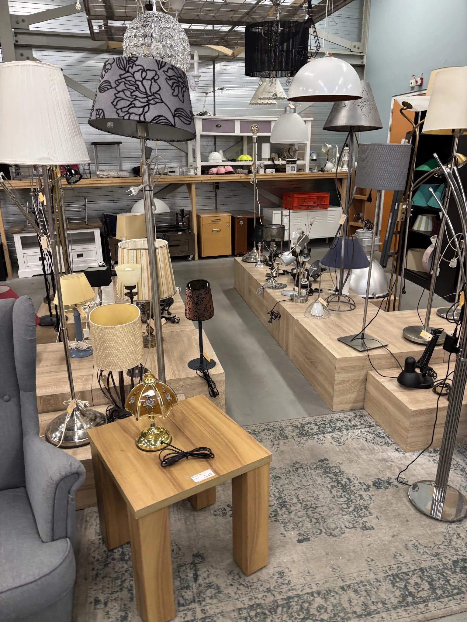 Winkelvloer van kringloopwinkel met houten werktafels, lampen, bureaustoelen en diverse spullen, goed verlicht interieur