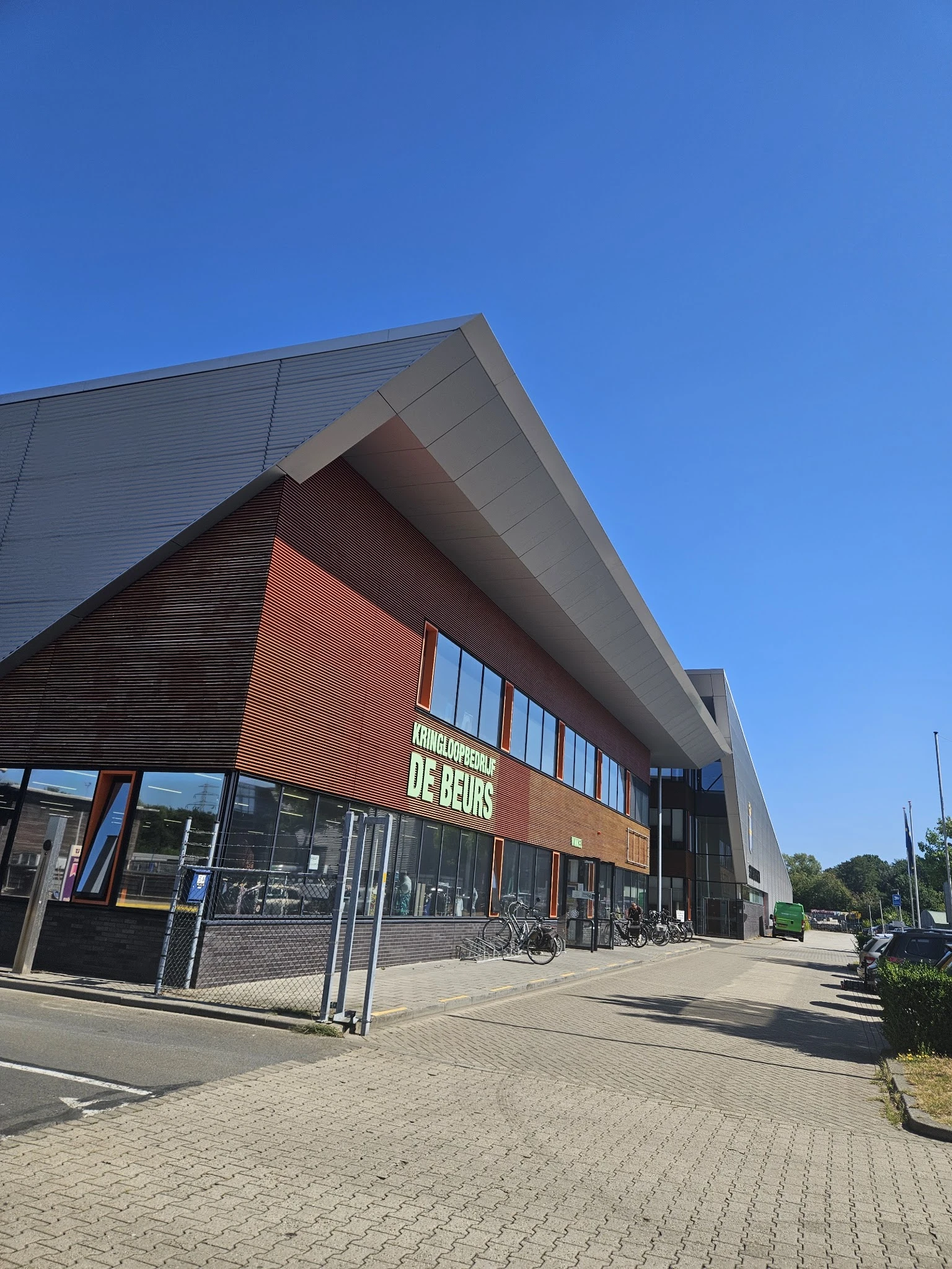 Moderne gevel van kringloopwinkel De Beurs met rode bakstenen en grijze dakconstructie, grote ramen en zichtbare winkelname.
