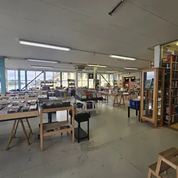 Ruime winkelruimte met tafels, stoelen, boekenkasten en diverse spullen uitgestald; grote ramen aan de zijkant