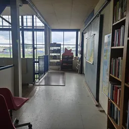 Gang in kringloopwinkel met boekenkasten aan rechts, ramen met uitzicht op landschap, rood zitmeubilair links
