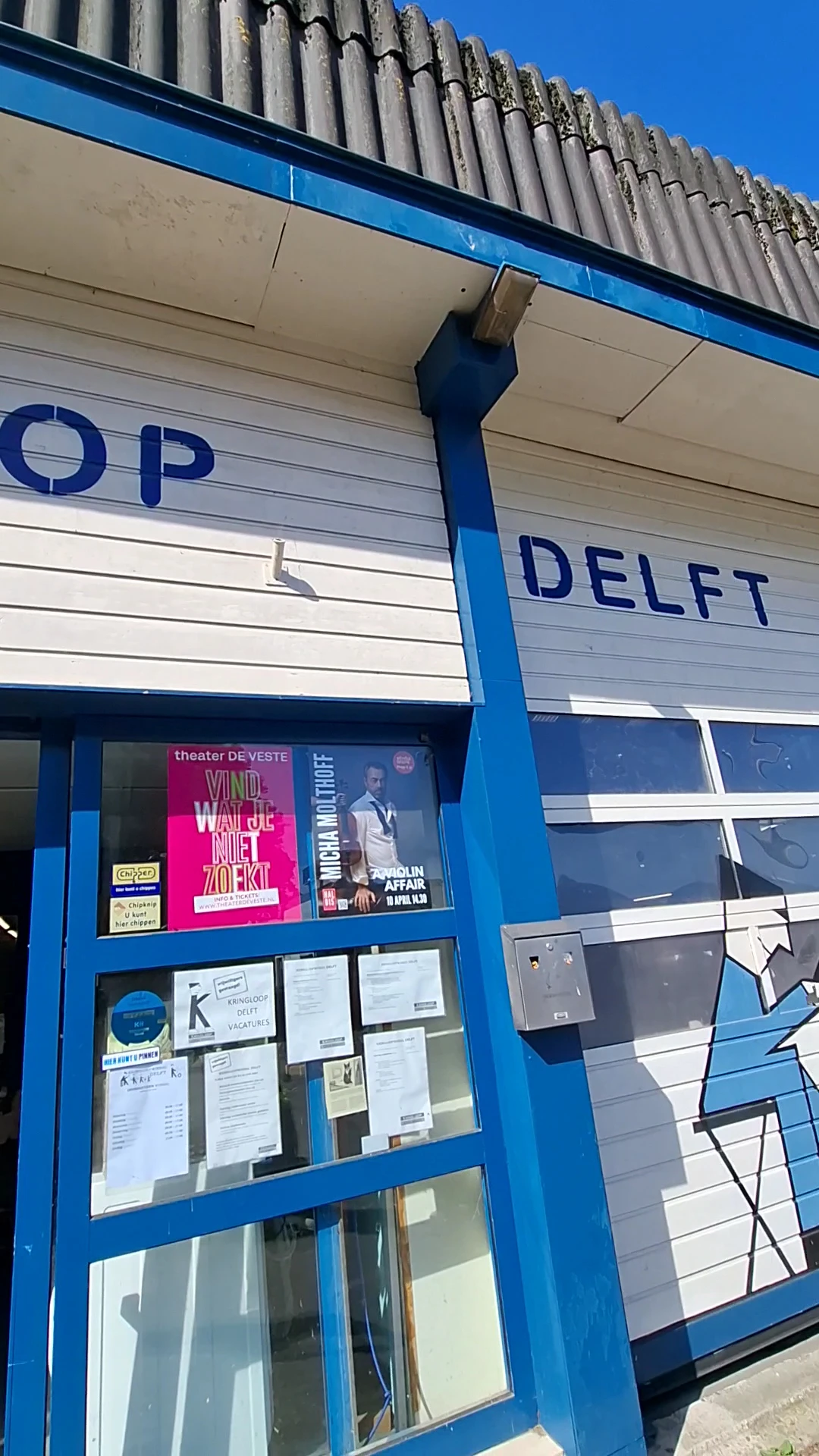 Blauwe gevel van kringloopwinkel met gedeeltelijk zichtbare naam DELFT en theater-poster, deel van pui zichtbaar