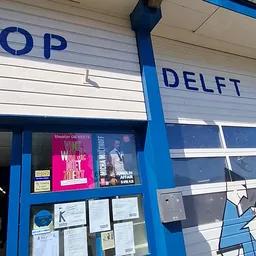 Blauwe gevel van kringloopwinkel met gedeeltelijk zichtbare naam DELFT en theater-poster, deel van pui zichtbaar