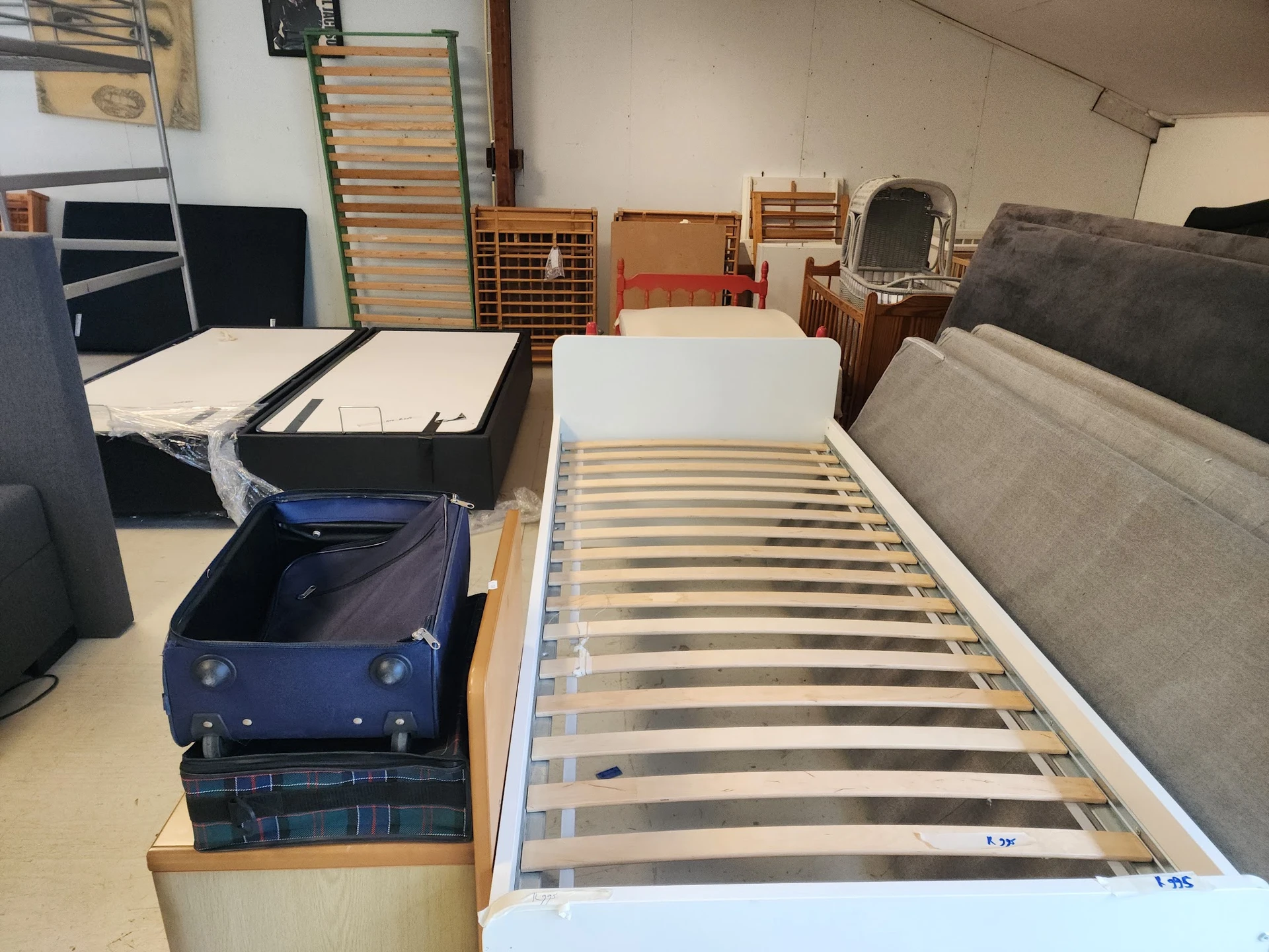 Uitgestalde bedden en matrassen in de kringloopwinkel, waaronder een wit eenpersoonsbed met lattenbodem op de voorgrond