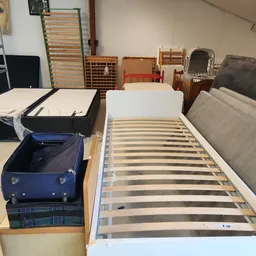 Uitgestalde bedden en matrassen in de kringloopwinkel, waaronder een wit eenpersoonsbed met lattenbodem op de voorgrond