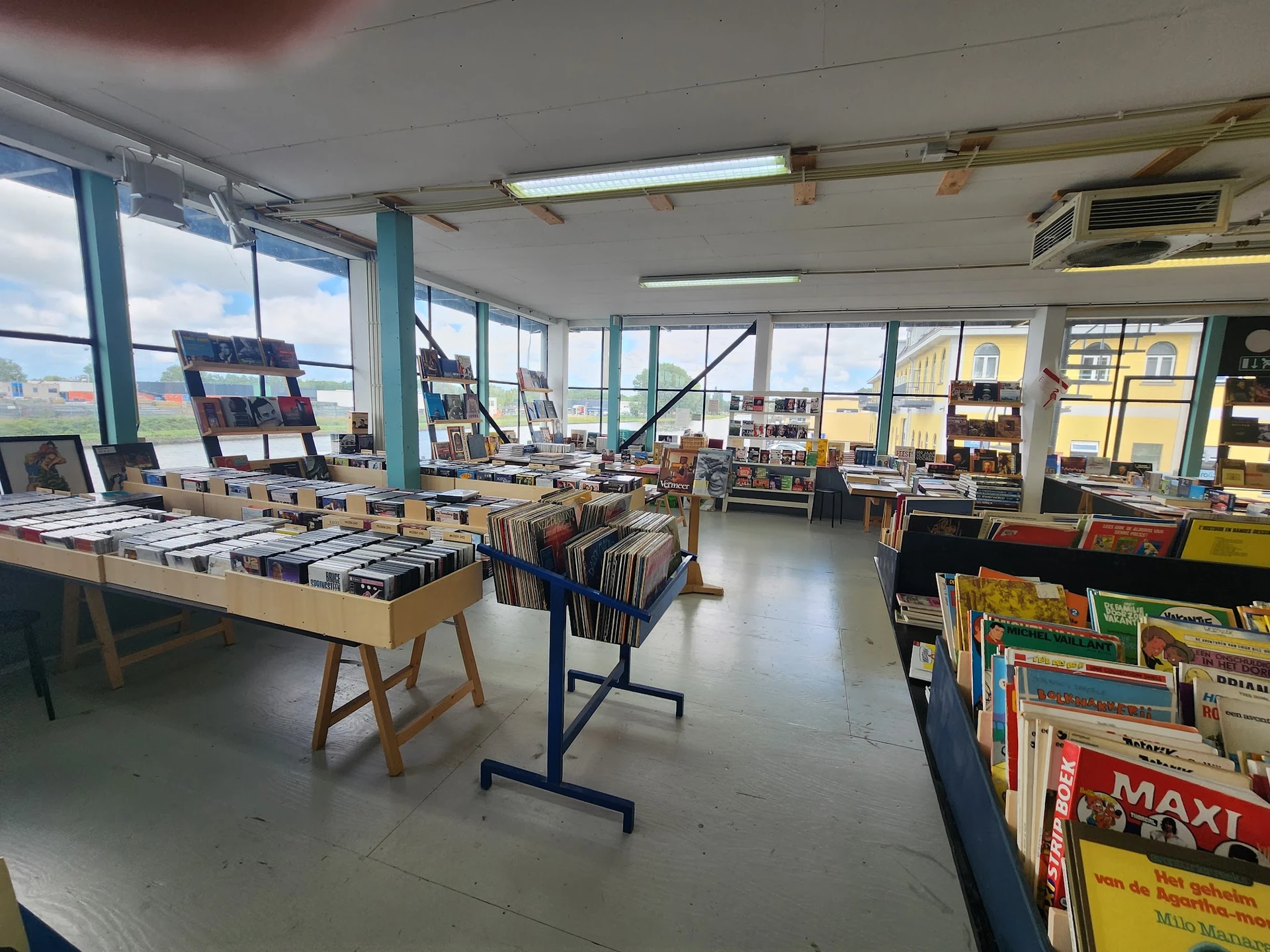 Ruime winkelvloer met tafels vol boeken, vinyl en media, grote ramen met uitzicht