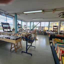Ruime winkelvloer met tafels vol boeken, vinyl en media, grote ramen met uitzicht