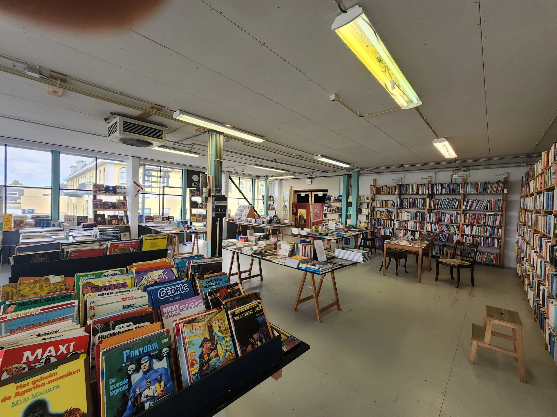Ruime winkelruimte met boekenkasten, speelgoed en diversen tegen de wanden, tafels in het midden, veel raam- en daglicht