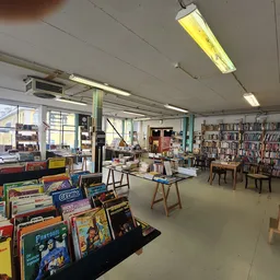 Ruime winkelruimte met boekenkasten, speelgoed en diversen tegen de wanden, tafels in het midden, veel raam- en daglicht