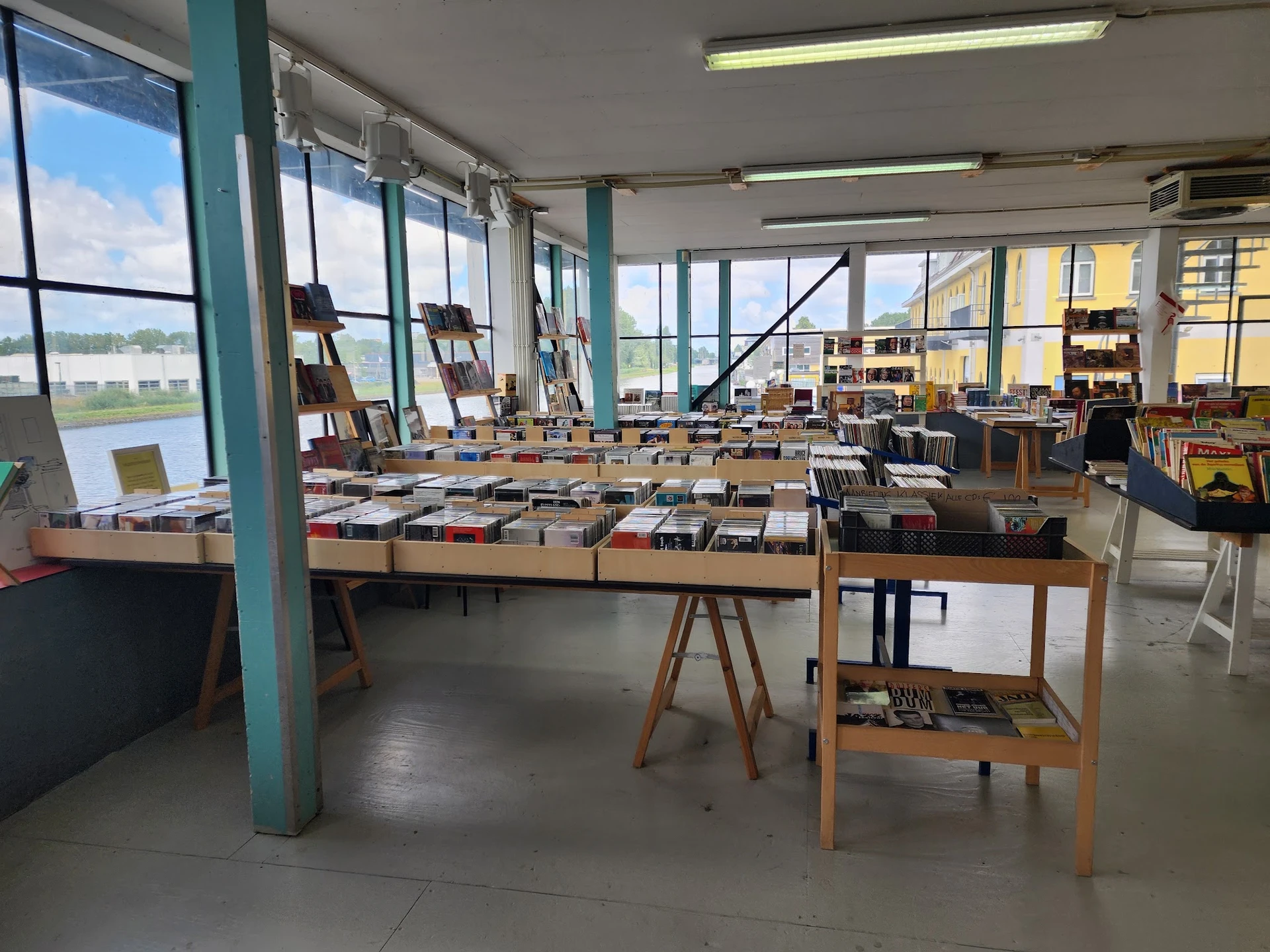 Ruime winkelvloer met tafels vol boeken, groene zuilen, grote ramen en boekenrekken in het achtergrond