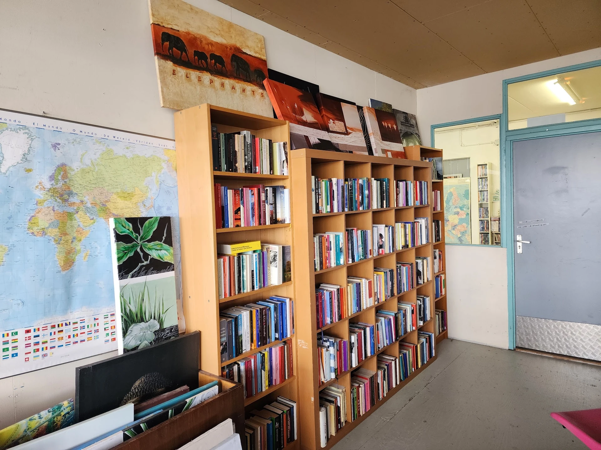 Boekenkasten vol boeken in een kringloopwinkel, met een wereldkaart en plantafbeelding aan de muur