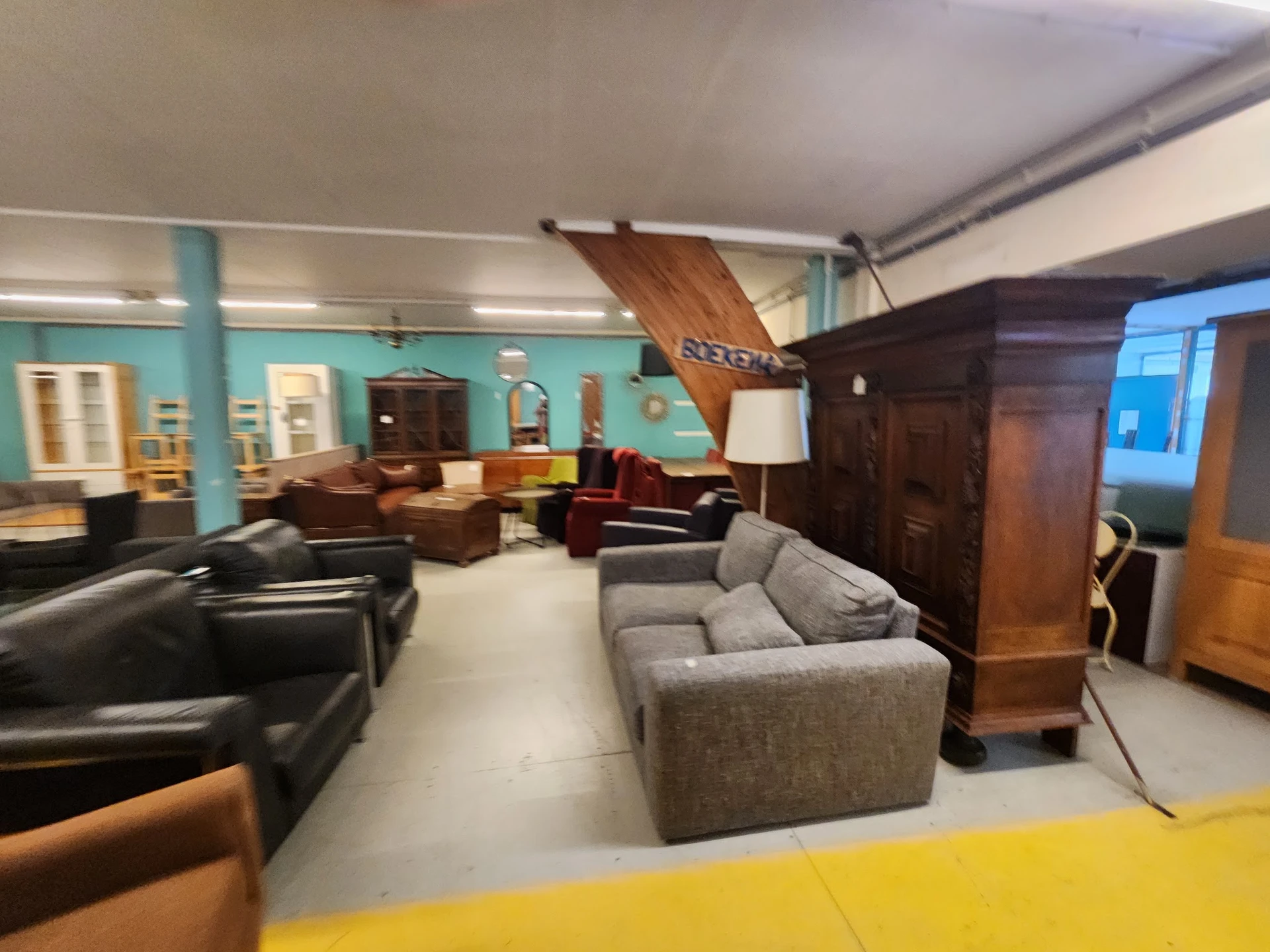 Ruim interieur van kringloopwinkel met meubels, zithoeken, houten boot aan plafond en turquoise wanden