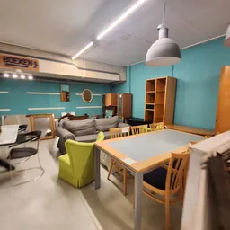 Ruime winkelruimte met turquoise wanden, houten meubels, tafel met stoelen en diverse tweedehands spullen