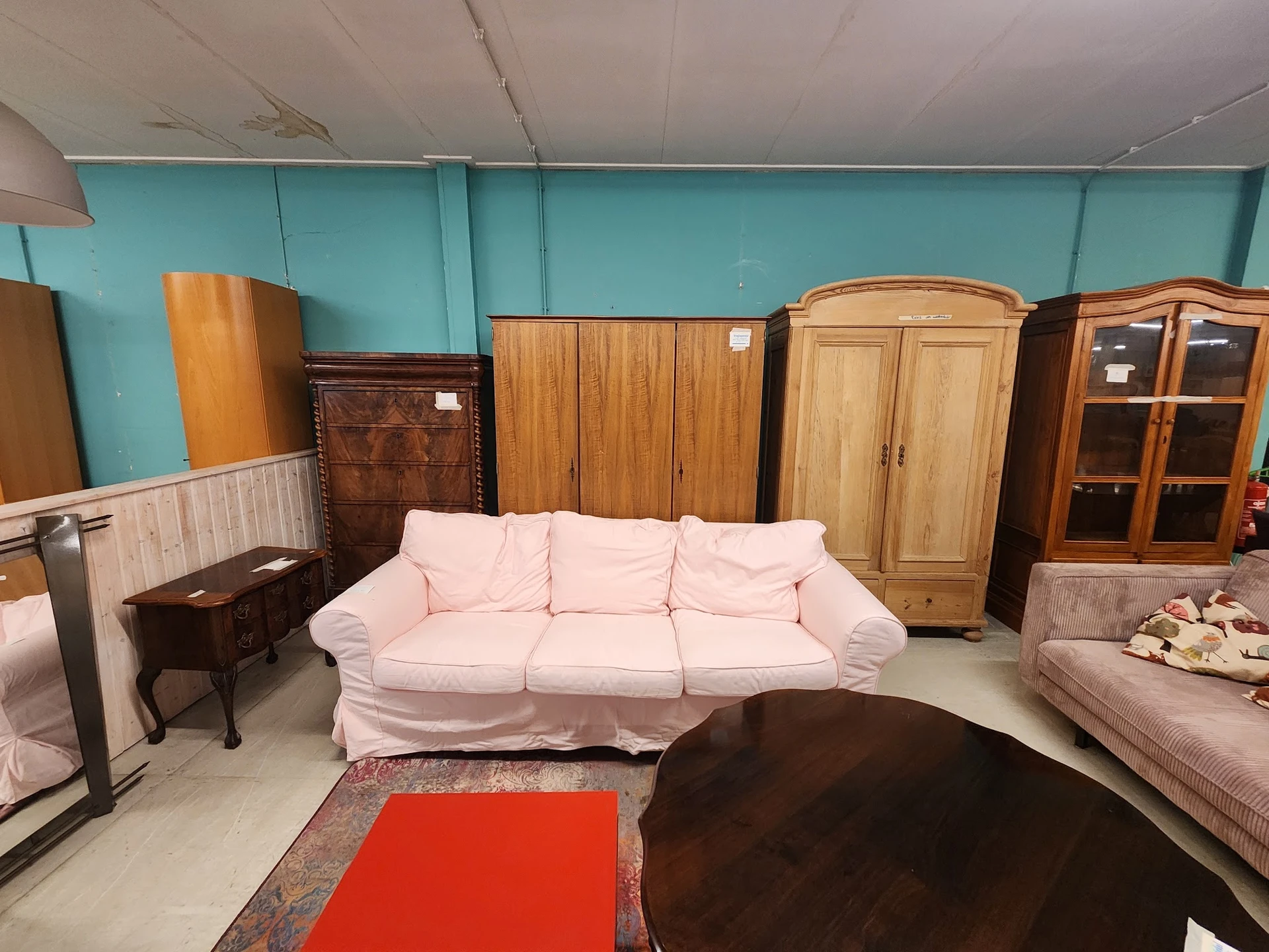 Winkelvloer met tweedehands meubels: roze bank, kasten, ladeblokken en andere meubelstukken tegen turquoise muur