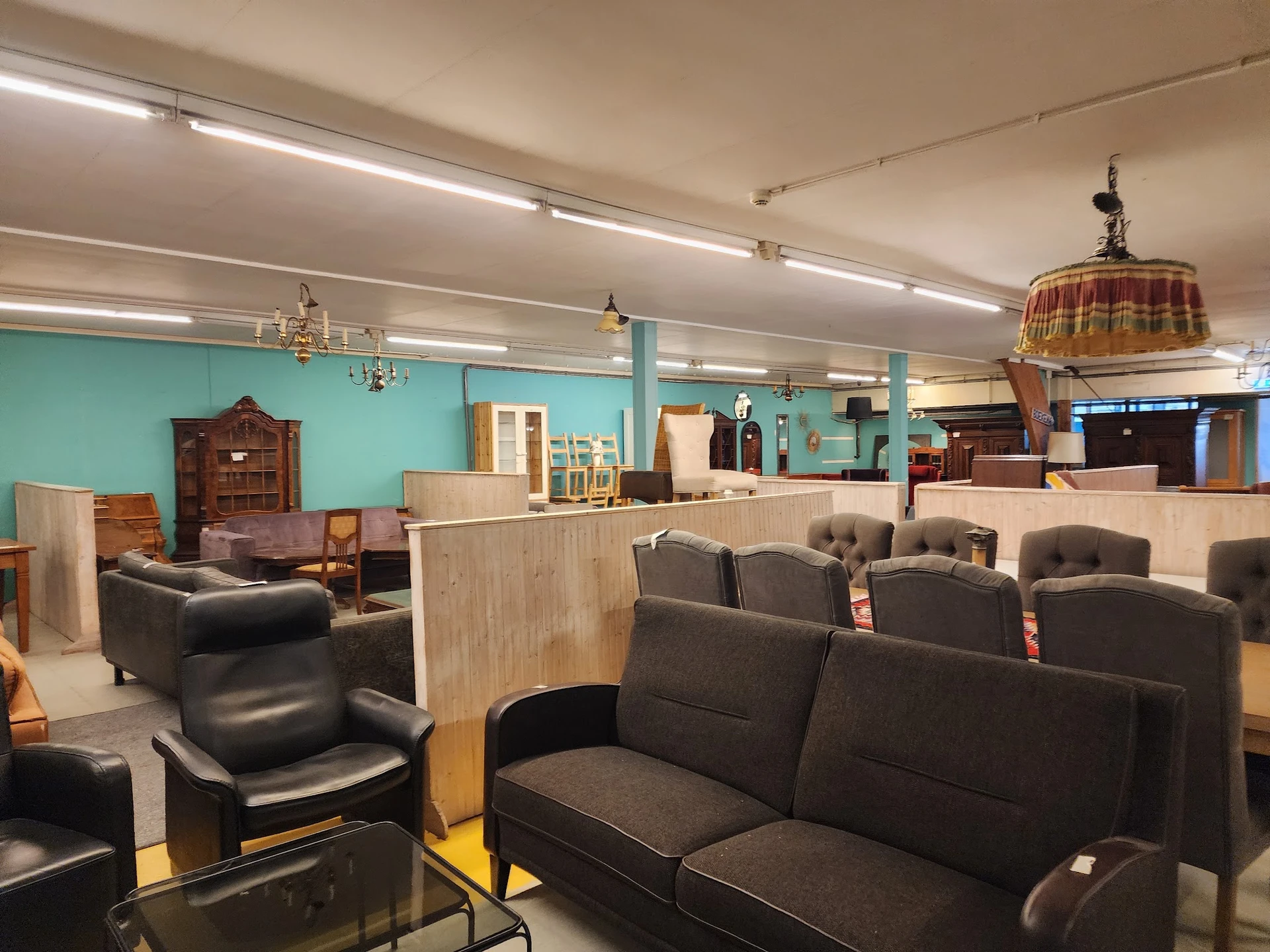 Winkelvloer met zitmeubels: zwarte fauteuils en banken, houten kasten en rekken, turquoise muren, fluorescerende verlichting