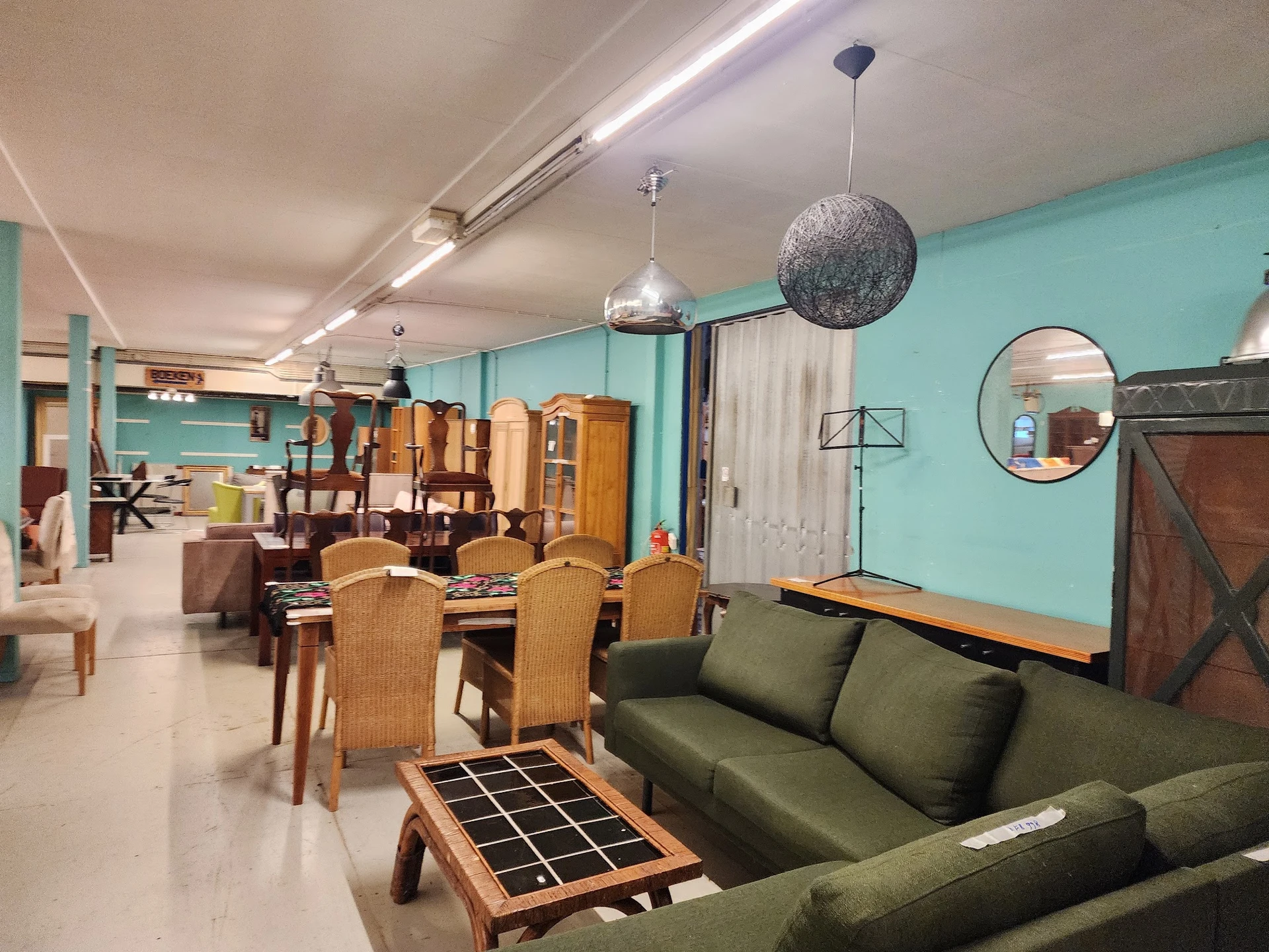 Woonkamer-achtig interieur met groen fluwelen bank, houten eettafel met rotan stoelen, en diverse meubels tegen turquoise mur
