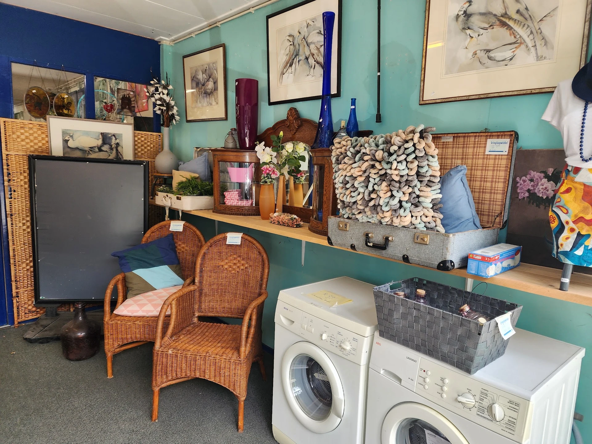 Afdeling met wasmachines, rieten stoelen, kunstwerk en decoratieve items op turquoise muur