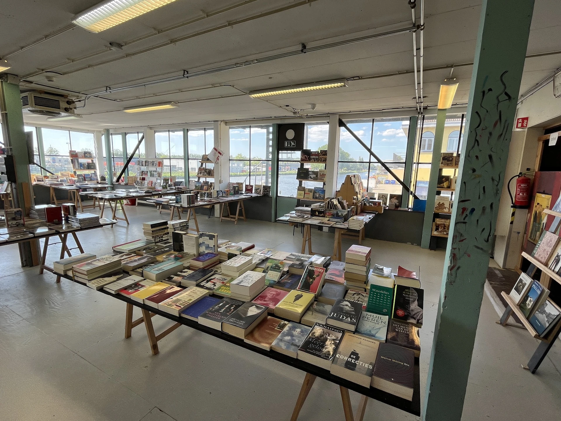 Lichte winkelruimte met lange tafel vol boeken en spullen, stellingen langs de wanden, grote ramen met uitzicht