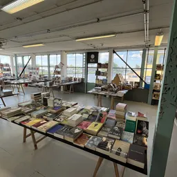 Lichte winkelruimte met lange tafel vol boeken en spullen, stellingen langs de wanden, grote ramen met uitzicht