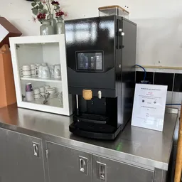 Zwarte koffieautomaat met displayscherm op een grijze werktafel, omgeven door andere winkelinventaris