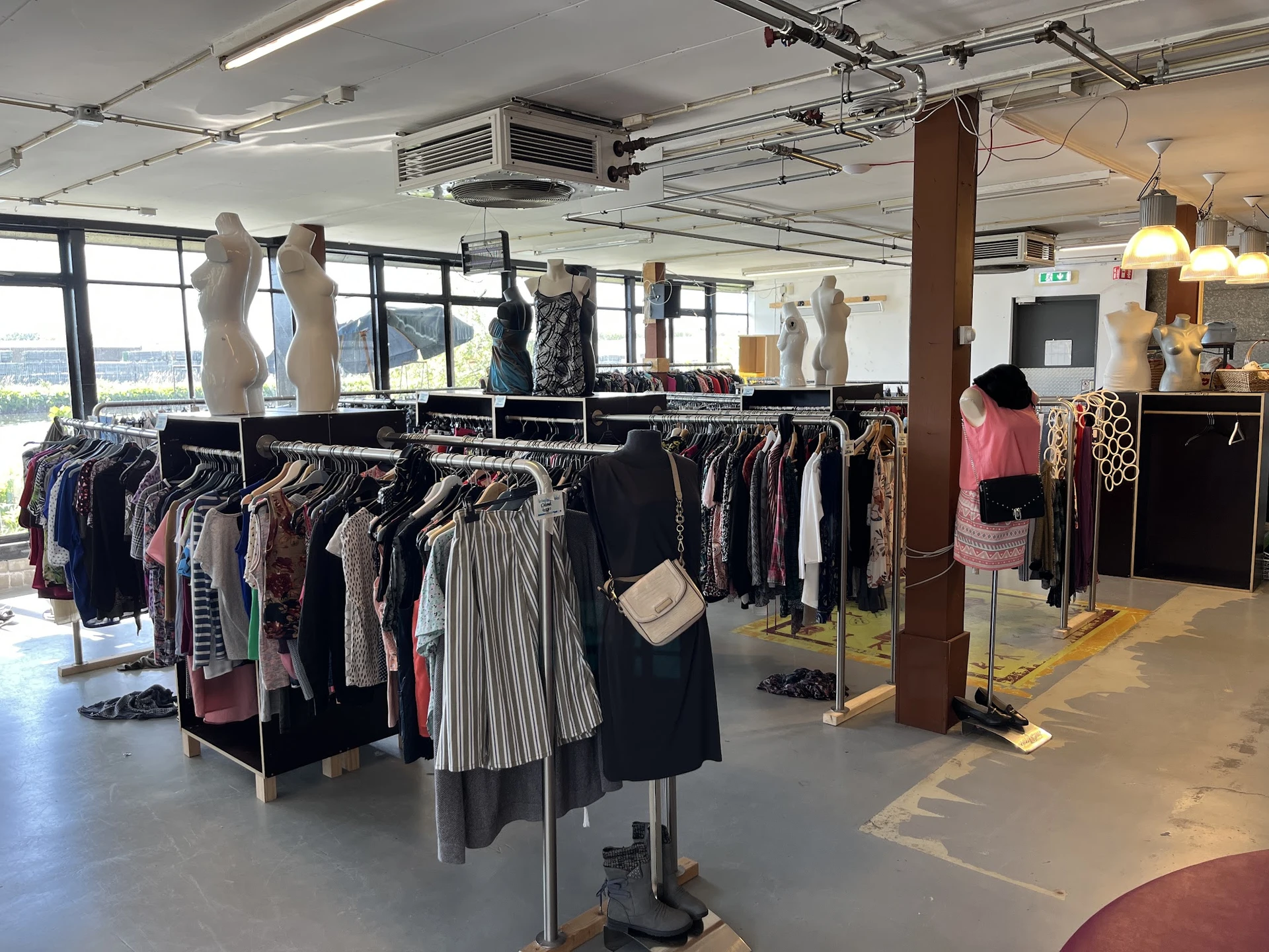 Rekken met tweedehands kleding, waaronder jurken, broeken en jassen, met etalagepoppen in een ruim kringloopwinkelbinnenruimt