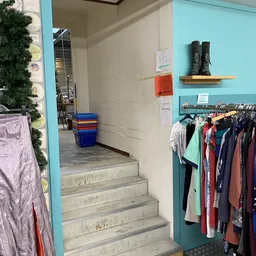 Trapopgang naar winkelruimte met rekken vol tweedehands kleding, schoenen op plank en kleurrijke kratten