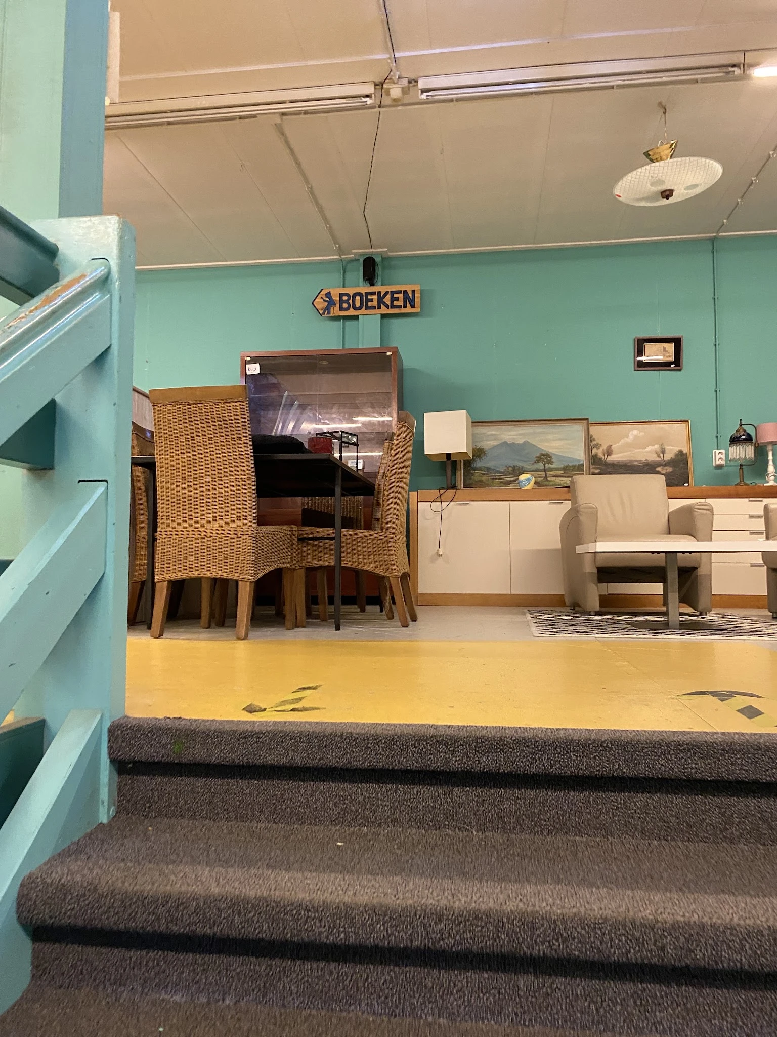 Uitgestalde meubels in de kringloopwinkel: rieten stoelen, houten tafels, kasten en beige stoelen tegen turquoise wand