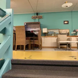 Uitgestalde meubels in de kringloopwinkel: rieten stoelen, houten tafels, kasten en beige stoelen tegen turquoise wand