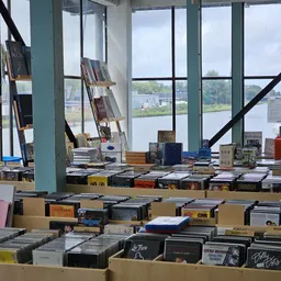 Tafels vol met tweedehands boeken en media, met stellages op de achtergrond bij grote ramen met uitzicht op water