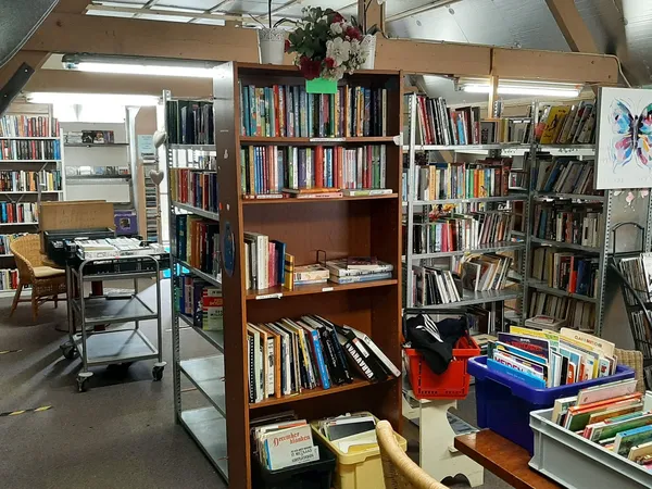 Binnenaanzicht van kringloopwinkel met boekenkasten, stellingen met boeken en cd's, werktafel en diverse spullen