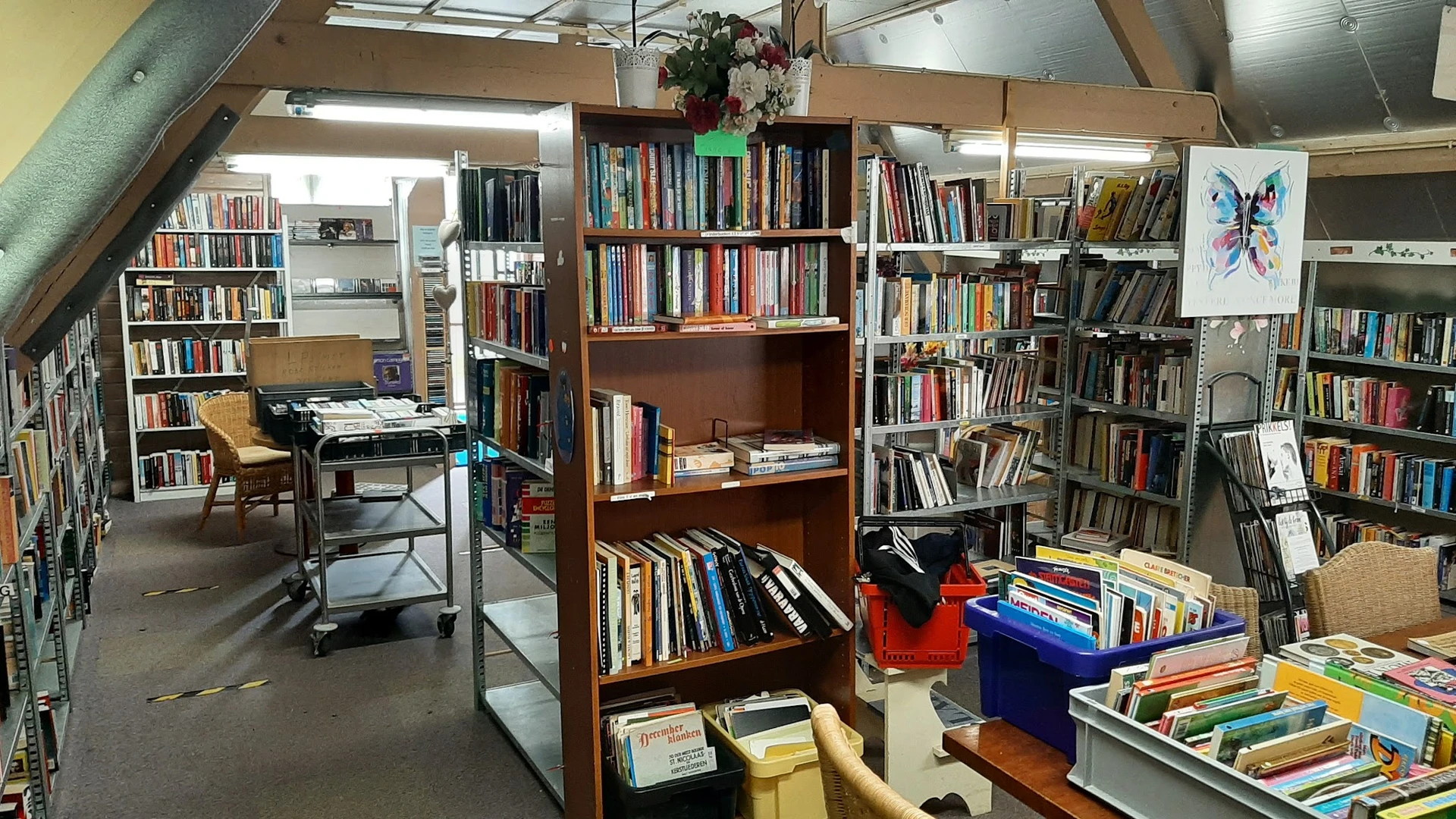 Binnenaanzicht van kringloopwinkel met boekenkasten, stellingen met boeken en cd's, werktafel en diverse spullen