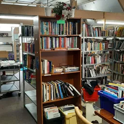 Binnenaanzicht van kringloopwinkel met boekenkasten, stellingen met boeken en cd's, werktafel en diverse spullen